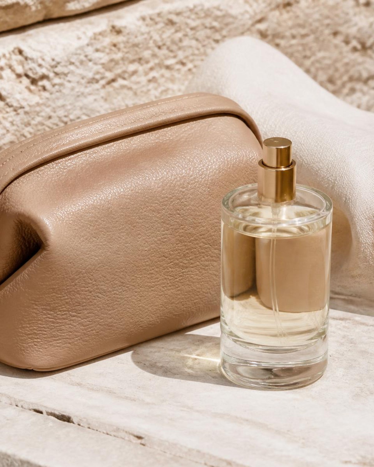 Unlabeled glass fragrance bottle and tan leather pouch on pale limestone with cream linen in warm sunlight